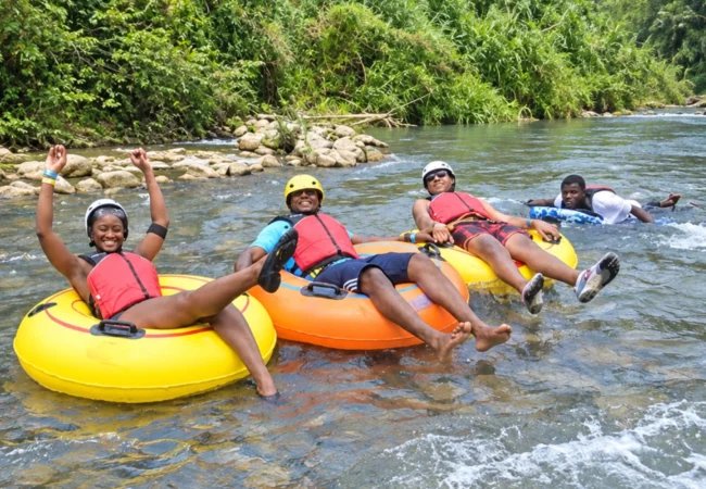 Three smiling tourists wearing safety helmets and red life jackets relaxing on yellow and orange inflatable tubes while enjoying a group river tubing adventure surrounded by lush tropical greenery in Ocho Rios Jamaica.