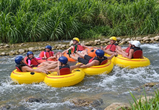 Group of tourists wearing colorful safety helmets and red life jackets enjoying river tubing on yellow and orange inflatable tubes through gentle rapids surrounded by lush tropical greenery in Ocho Rios Jamaica.